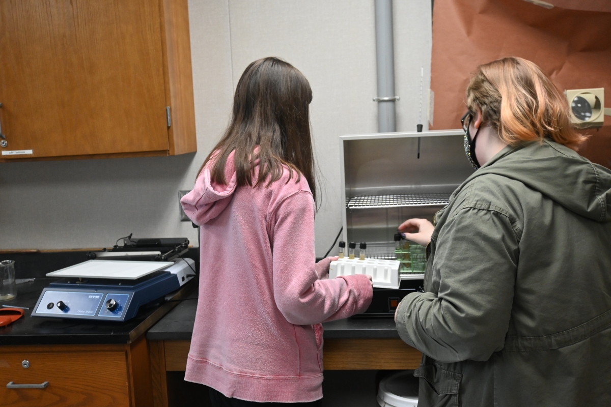 Two high school students stand near a lab incubator